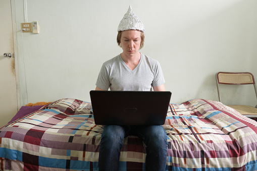 Portrait of young handsome man wearing tin foil hat as conspiracy theory concept in the bedroom at home