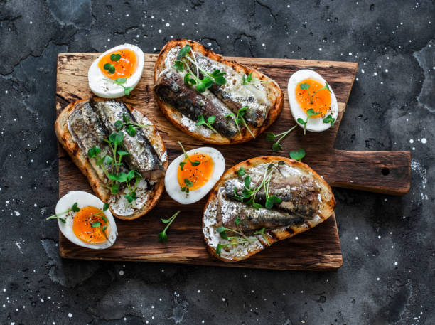 delicioso aperitivo, tapas - huevos cocidos suaves y sándwiches de sardinas enlatadas en una tabla de cortar sobre un fondo oscuro, vista superior - salad breakfast cooked eggs fotografías e imágenes de stock