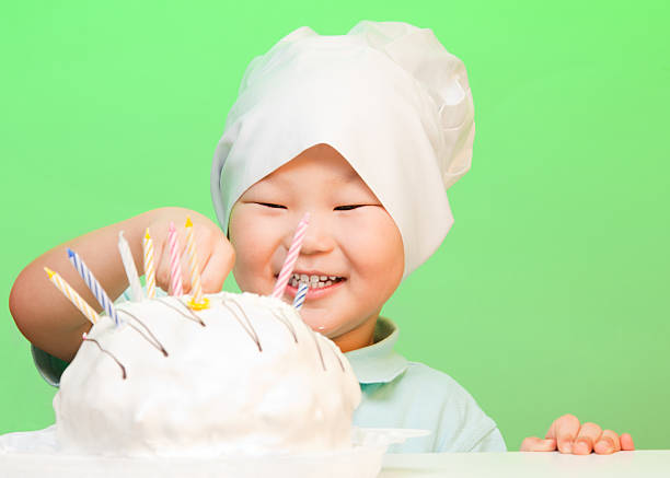 Chinese boy putting candles in birthday cake, smiling and wearing cooks hat