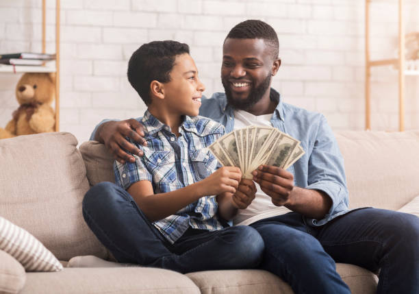 glücklicher afro papa und preteen sohn mit dollar bargeld zu hause - currency savings coin counting stock-fotos und bilder