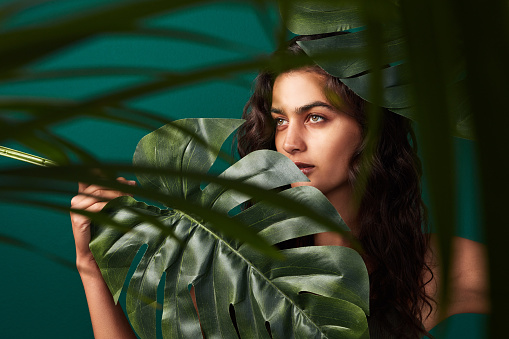 Cropped shot of a beautiful young woman posing with monstera leaves against a green background