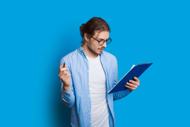 istock Caucasian bearded boy with long hair is looking in a notebook through eyeglasses and posing on a blue wall 1209194875