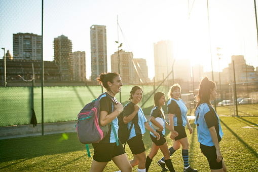 Side view with lens flare of Hispanic female football team walking onto sports field with gear for afternoon practice.