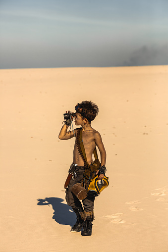 Post apocalyptic boy outdoors in the desert. People in nuclear post-apocalypse. Life after doomsday concept. Desert and dead wasteland copy space background. A young man walking with telescope, knife, gas mask, mug looking for someone.