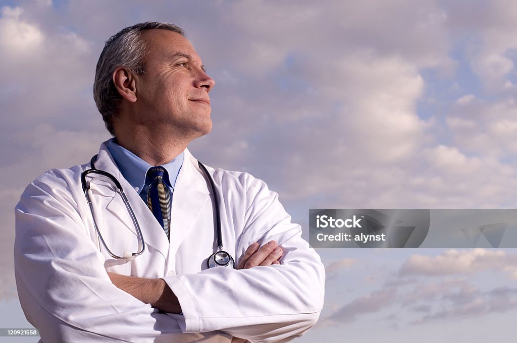 Stock Photo of a Confident Medical Doctor with Copy Space Stock photo of a confident, medical, male doctor standing with his arms folded and looks off to the distance with a smile, against a partially cloudy sky. Copy space. Doctor Stock Photo