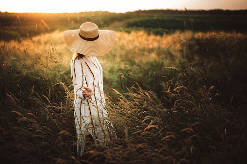 Woman in rustic dress and hat walking in wildflowers and herbs in sunset golden light in summer meadow. Stylish girl enjoying evening in countryside. Rural slow life. Copy space