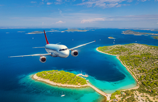 Airplane is flying over small islands and sea at sunny day in summer. Aerial view of passenger airplane, tropical seashore, mountains with green trees, sky and blue water. Top view of aircraft. Travel