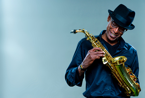 Musician with saxophone  against a grey backdrop. Man looking down with a smile, wearing a black hat and shirt