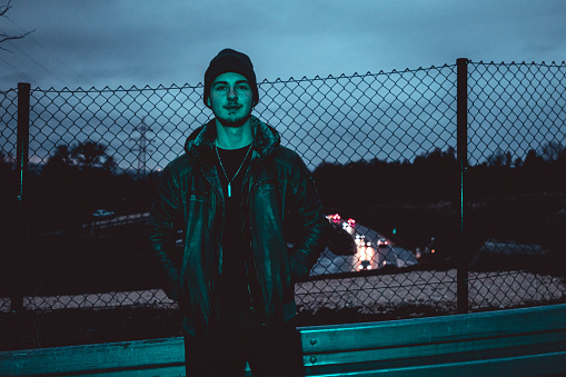 Young teenage man standing in front of urban highway fence at night, looking towards the camera. Youth culture teenage lifestyle portrait.