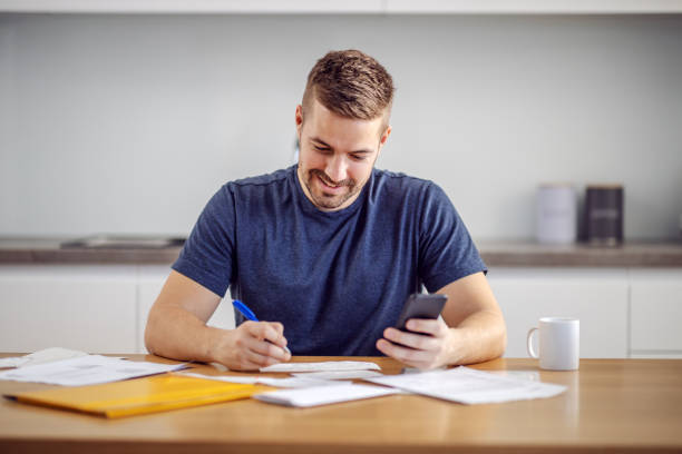 Young handsome smiling bearded man sitting at home, calculating budget and filling in bills. Young handsome smiling bearded man sitting at home, calculating budget and filling in bills. beak stock pictures, royalty-free photos & images