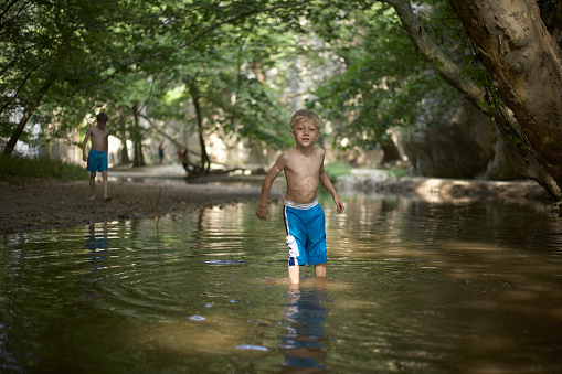 Little boy aged 5 or 6 or 7 standing in shallow water in forest in mediterranean country in summer time sunny relaxed mood teenage boy in background
