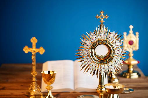 Catholic symbols composition. The Cross, monstrance,  Holy Bible and golden chalice on the altar. Blue background.
