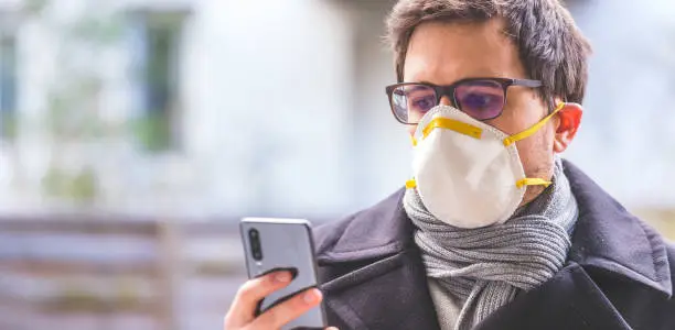 Young business man outdoors with a breathing mask and smartphone. Flue and corona season.