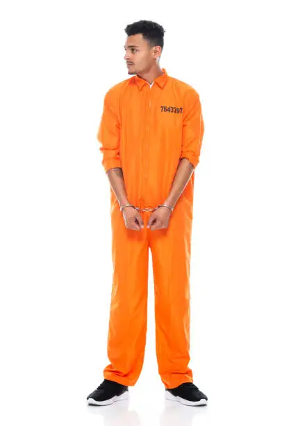 Full length of aged 20-29 years old generation z young male criminal standing in front of white background wearing uniform who is depressed and using handcuffs