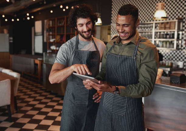 istock Cheerful cafe partners wearing apron standing inside a coffee shop looking on their touchscreen 1209263620