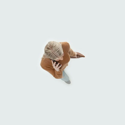 Aerial view of aged 20-29 years old who is beautiful with blond hair caucasian female walking in front of white background wearing warm clothing who is talking and holding purse and using mobile phone
