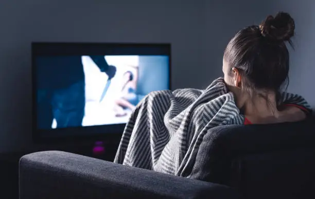 Photo of Scary horror movie on tv. Scared woman watching stream service hiding under blanket on couch at night. Sleepless person streaming series or film on television.