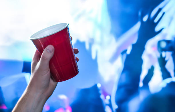 coupe rouge de partie dans la main dans la boîte de nuit, bar ou événement d’étudiant d’université. tasse de bière en plastique. les gens ayant l’amusement dans les lumières bleues de disco de boîte de nuit sur la piste de danse - disposable cup red beer plastic photos et images de collection