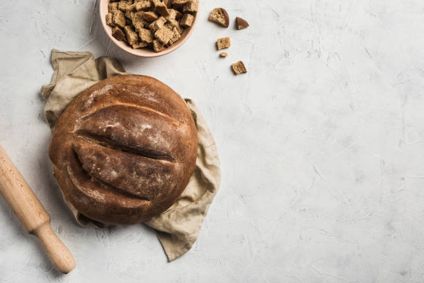 A round loaf of bread lies on a grey linen napkin with breadcrumbs in a bowl and a rolling pin for dough on a light background.