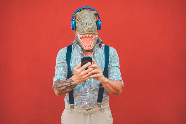 tätowierter mann mit t-rex-maske mit smartphone beim hören musik - crazy senior kerl auswahl playlist aus handy-app - technologie-trends und wahnsinn kostüm-konzept - fokus auf gesicht - headphones music mobile phone tattoo stock-fotos und bilder