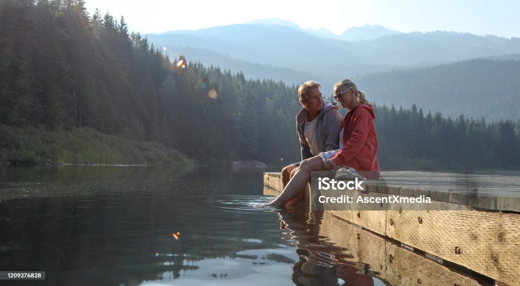 Mature couple relax on wooden pier, looks out across lake Lost Lake, Whistler, BC Mature Couple Stock Photo