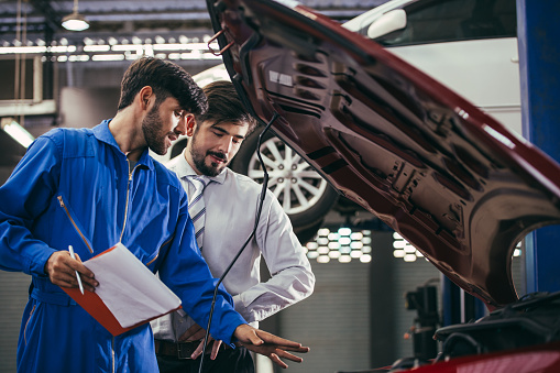 automotive maintenance mechanic young man explain car condition to male customer in garage at auto repair shop, technician pointing needed on vehicle checklist document, after service concept