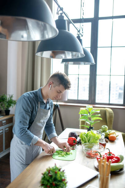 istock Blond-haired man in wireless headphones and apron preparing salad. 1209290378