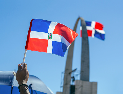 A hand holding the Dominican flag with the monument in the background