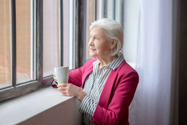 istock Grey-haired smiling lady standing at the window and thinking 1209299619