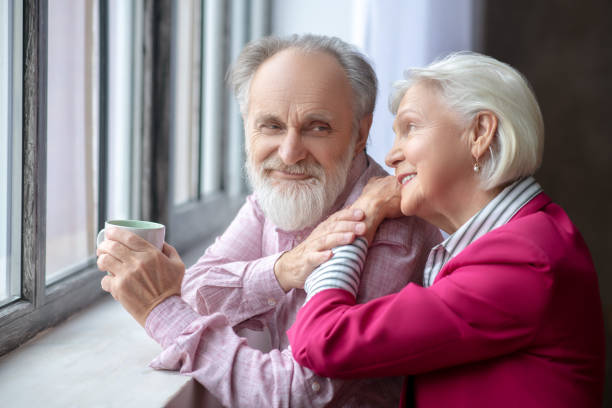 istock Elderly grey-haired couple standing near the window looking happy 1209299625