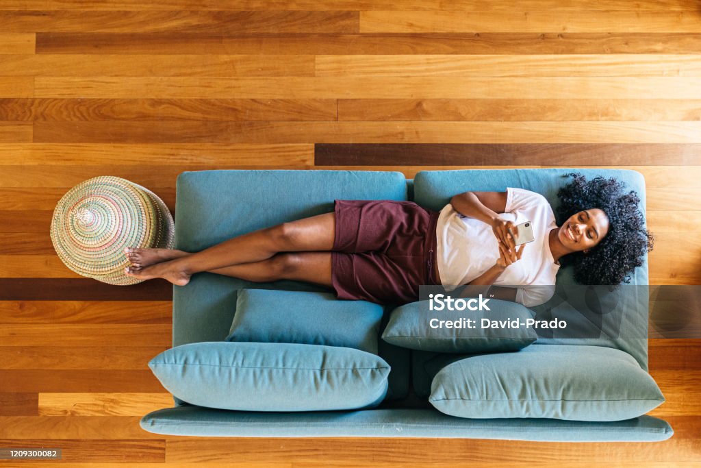 Black woman chatting on smartphone lying on couch at home Top view of African American lady in casual wear resting on blue sofa and smiling while surfing on mobile phone at home Sofa Stock Photo