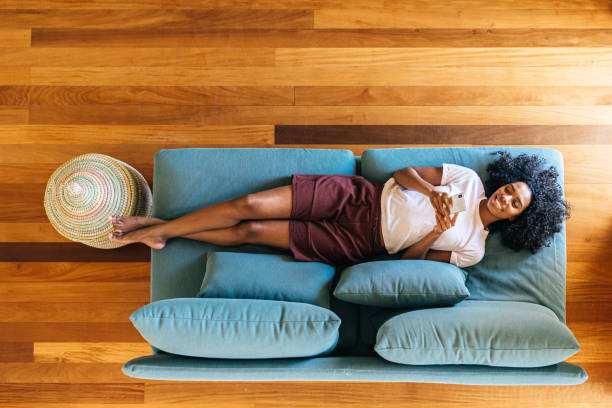 Black woman chatting on smartphone lying on couch at home Top view of African American lady in casual wear resting on blue sofa and smiling while surfing on mobile phone at home lifestyle people stock pictures, royalty-free photos & images
