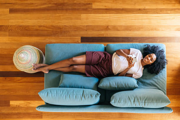 black woman chatting on smartphone lying on couch at home - fotos de aconchegante imagens e fotografias de stock