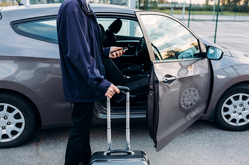 Unrecognizable man standing at car with suitcase and holding cell phone