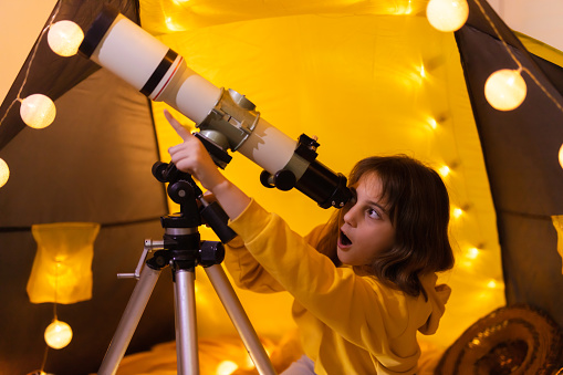 Small girl using telescope at home living room in a tent.