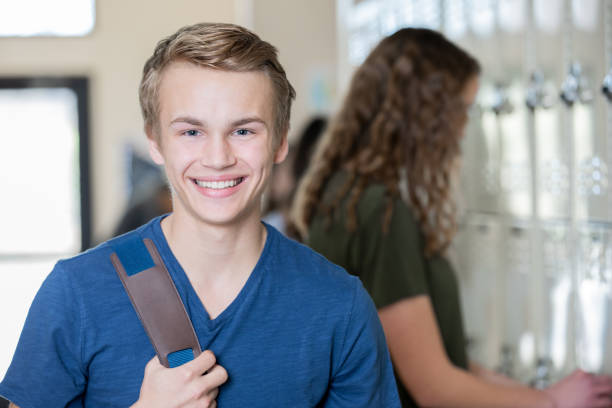 istock Teenage boy smiles while looking at camera in high school hallway by lockers 1209311994
