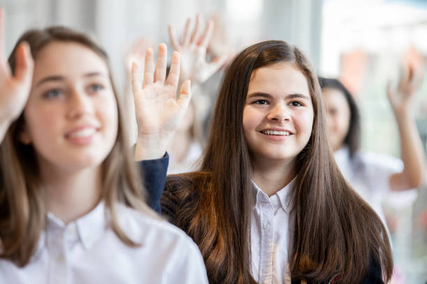 Female high school student smiles while raising hand in classroom Female high school student smiles while raising hand in classroom teenage high school girl raising hand during class stock pictures, royalty-free photos & images