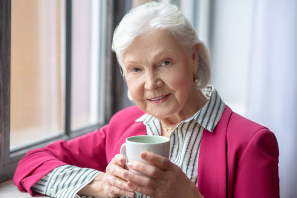 istock Grey-haired smiling lady standing near the window with a cup in her hand 1209350961