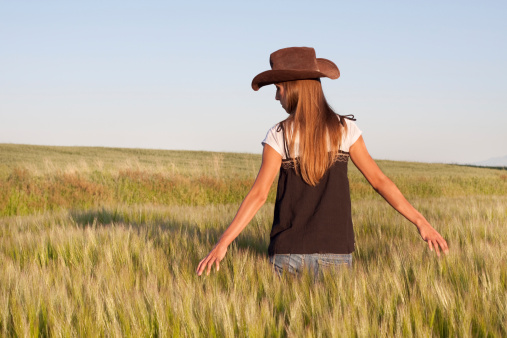 Anonymous woman with long hair and wearing cowboy hat, walking through grain field.