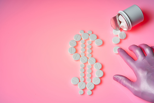 White pills are in the form of a dollar sign on a pink background, a bottle with medicines, a hand in a glove moves the pill. Pharmaceutical business concept
