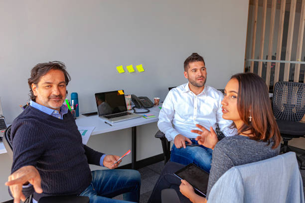 Two Millennial Generation Latin American White Collar Workers Dressed In Casual Clothes Have An Impromptu Meeting With Their Manager In A Small Office