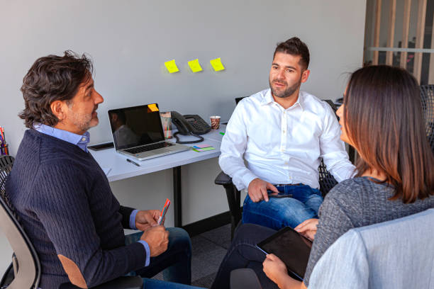 Two Millennial Generation Latin American White Collar Workers Dressed In Casual Clothes Have An Impromptu Meeting With Their Manager In A Small Office