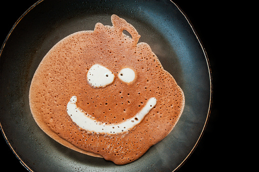 A pancake made of liquid chocolate dough with bubbles is fried in a black pan with drops of fat and oil on an induction cooker. Funny food with a face image.