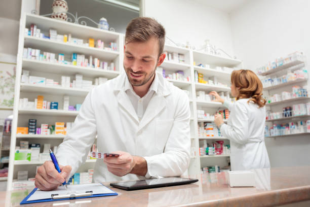 farmacéutico joven de pie detrás del mostrador en una farmacia, escribiendo en un portapapeles - receta instrucciones fotos fotografías e imágenes de stock