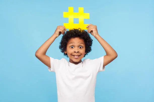 Photo of Funny excited surprised little boy with curly hair in T-shirt holding big yellow hashtag symbol over his head