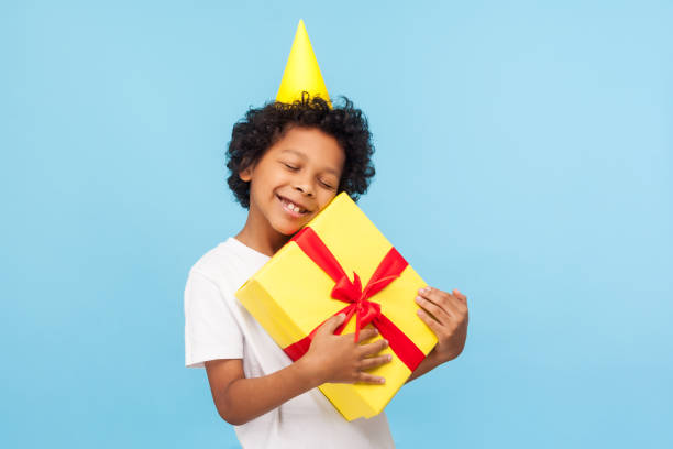 istock Amusing happy pleased little boy with party cone on head embracing gift box and closing eyes in pleasure 1209423755
