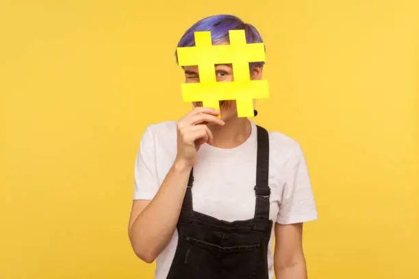 Hashtag, interesting media trends. Portrait of cheerful young woman with violet short hair in denim overalls looking at camera through big hash sign with curious eyes. yellow background, studio shot