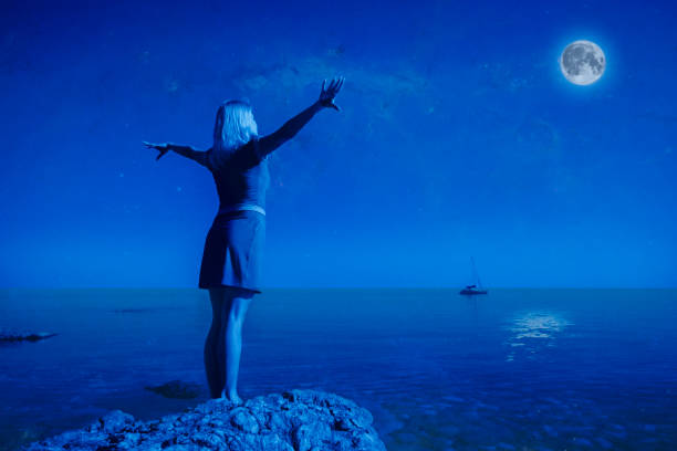 Rear view unidentified young happy woman stands on a stone raise Hands up Looking at the big moon and calm clear sea water against the backdrop of landscape and clear night sky. Full moon concept