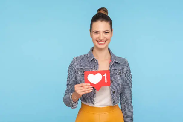 Photo of Portrait of joyful beautiful fashionably dressed woman with hair bun holding social media heart icon, network one Like button