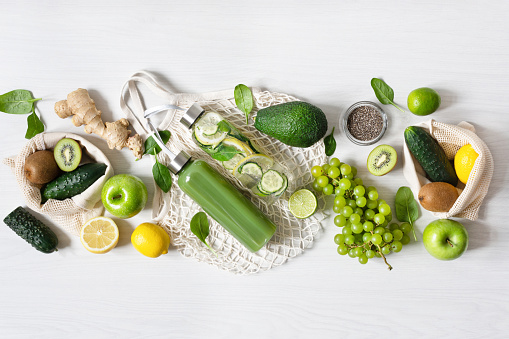 Various green fruits and vegetables with bottles of smoothie and infused water on white wooden table top view. Eco friendly shopping concept. Detox diet.